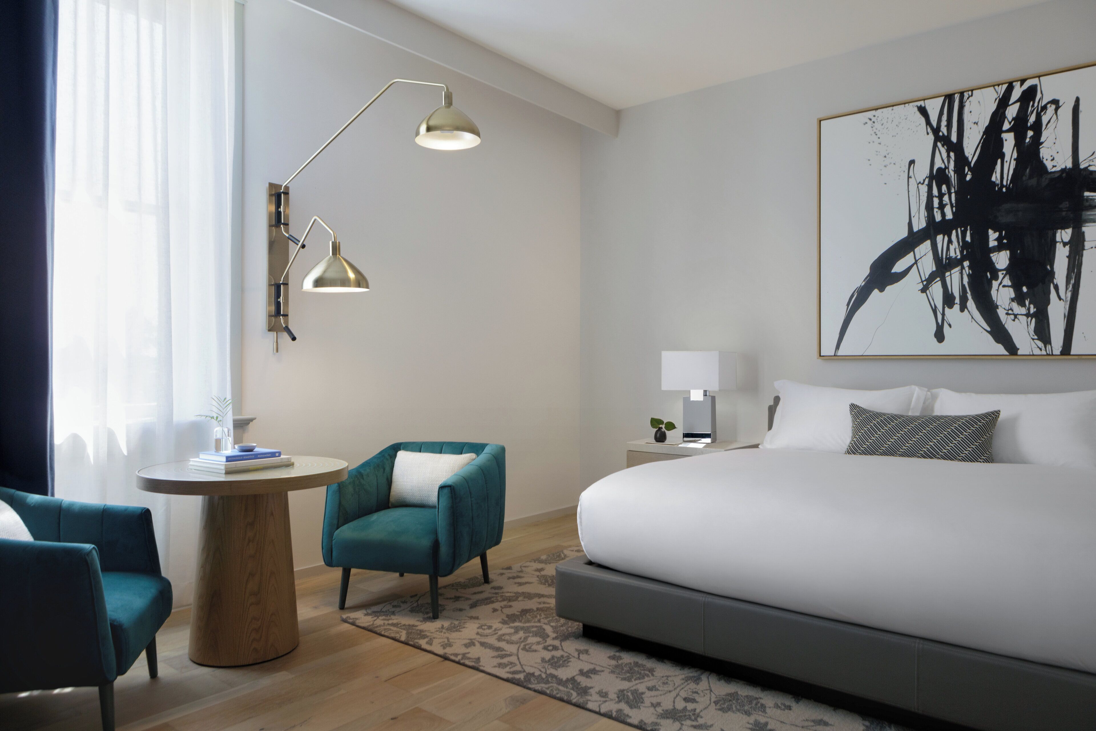 Una habitació d'hotel moderna amb un llit gran, dues butaques, una taula rodona petita, una làmpada i un quadre abstracte de grans dimensions a la paret.
