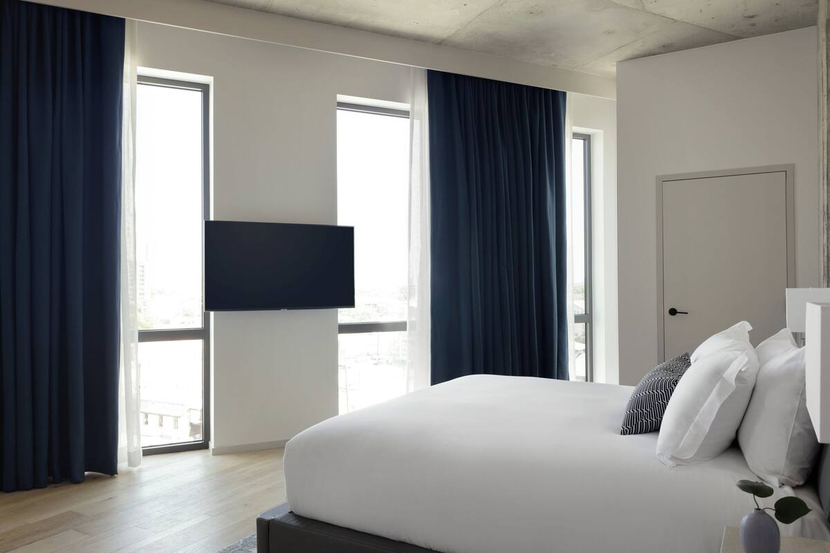 A modern hotel room with a large bed, dark curtains, a flat-screen TV, and a view of the city through large windows.