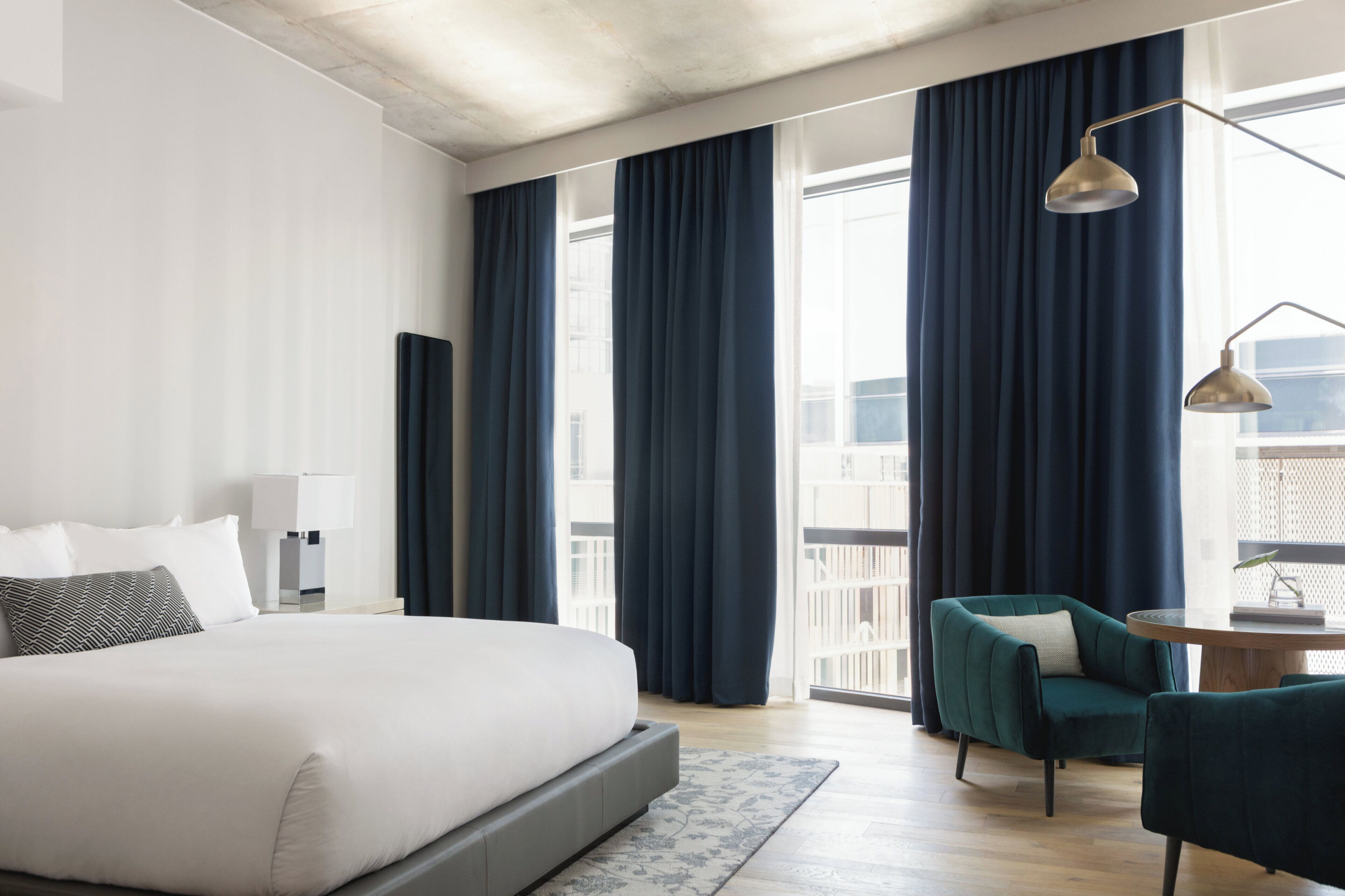 Ein modernes Hotelzimmer mit einem großen Bett, einem Schreibtisch mit Lampe, einem Sessel und Blick auf die Stadt durch große Fenster.