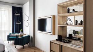 A modern living room with a sofa, a coffee table, and a TV. There is a bookshelf with decorative items and a window with curtains.