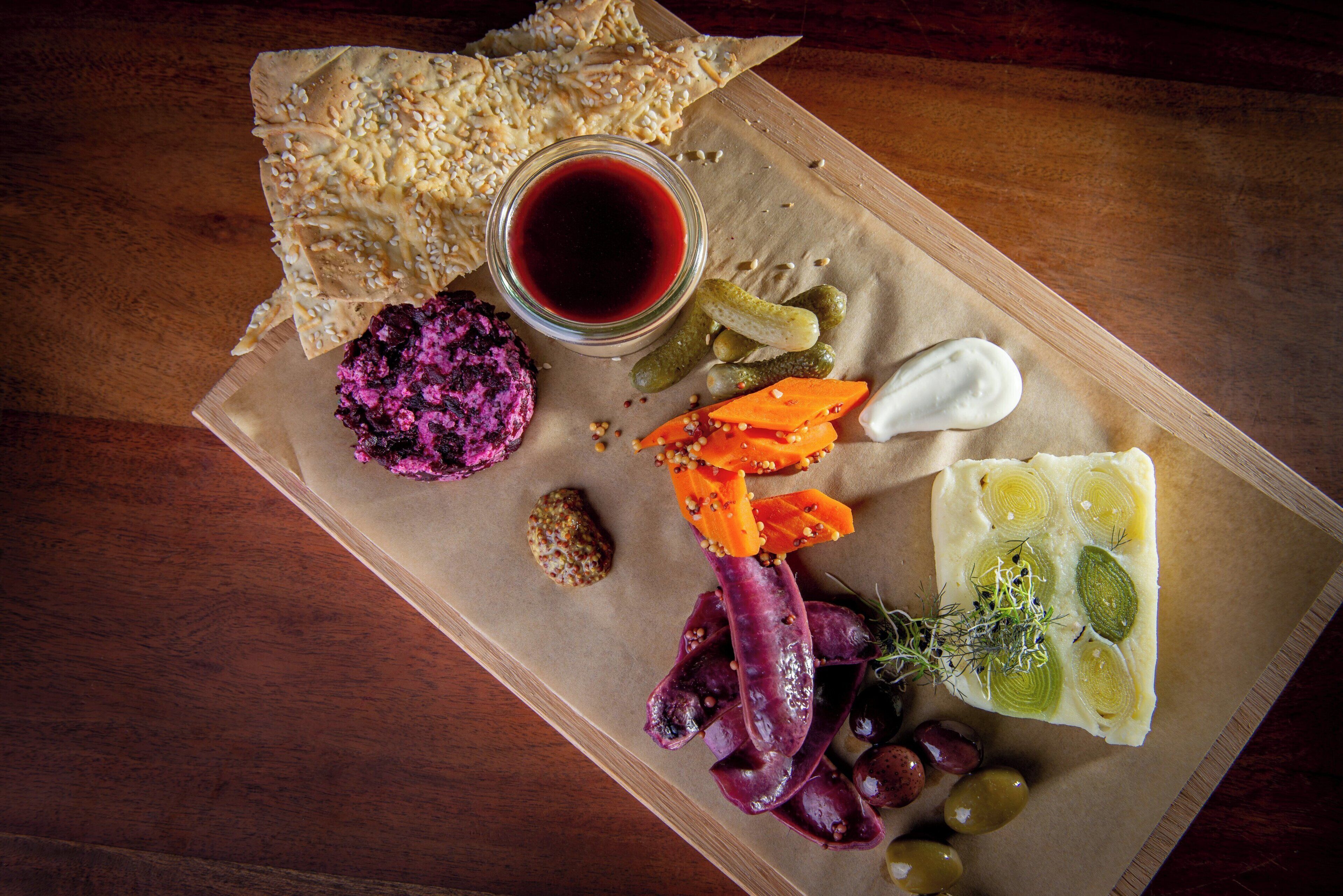 A wooden board with various food items including pickles, a beetroot dish, olives, and a cheese board with herbs.