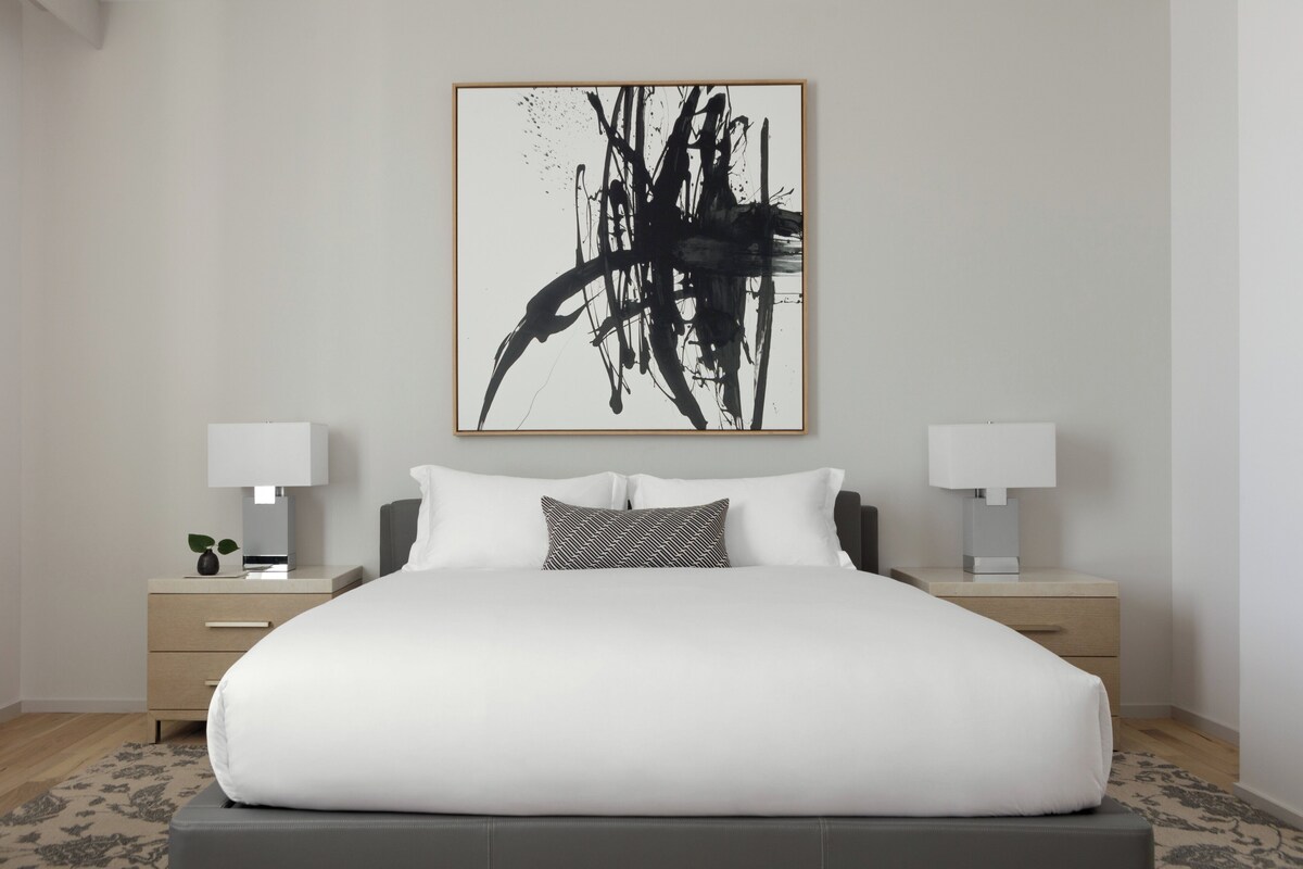 A modern bedroom with a large bed, two bedside tables with lamps, and a large abstract painting on the wall.