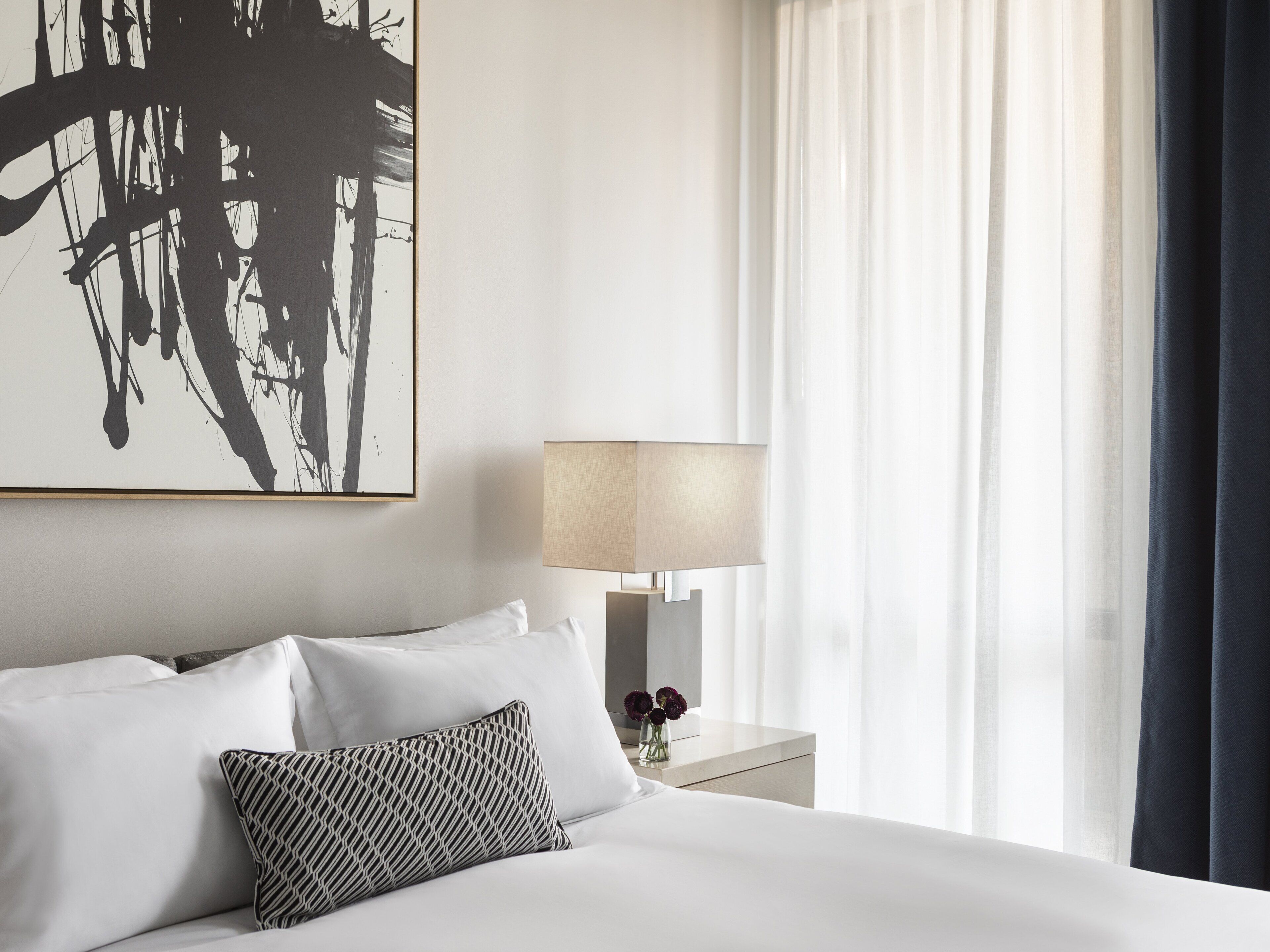 Une chambre d’hôtel avec un grand tableau abstrait, un lit recouvert de draps blancs, un oreiller à motifs, une lampe de chevet et des rideaux transparents.