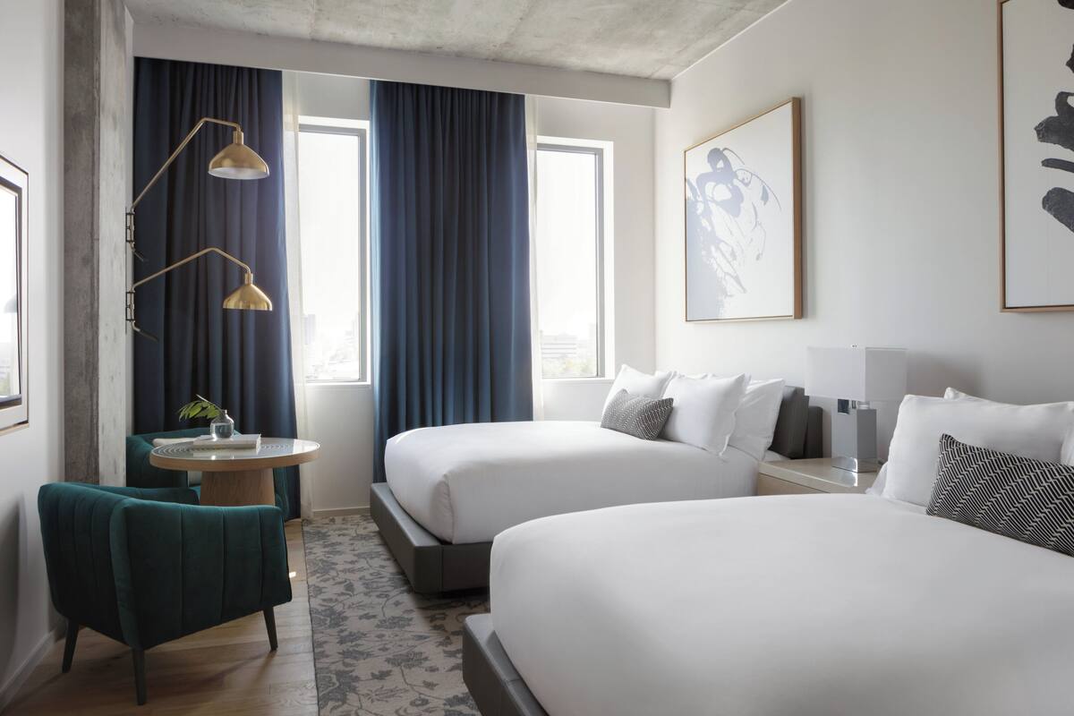 A hotel room with two beds, a desk, and a chair. There are two windows with curtains, a floor lamp, and artwork on the walls.