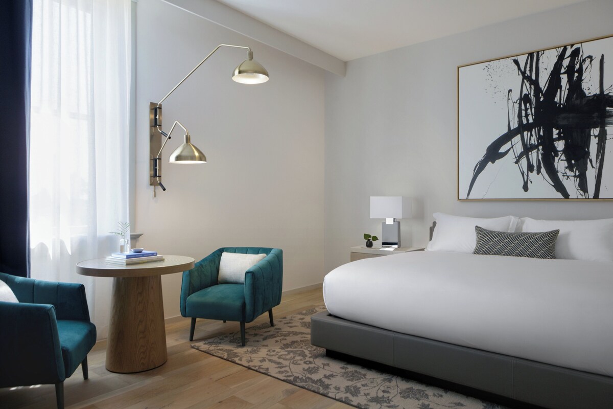 Una habitación de hotel moderna con una cama grande, un escritorio, una silla, una lámpara y un cuadro en la pared.