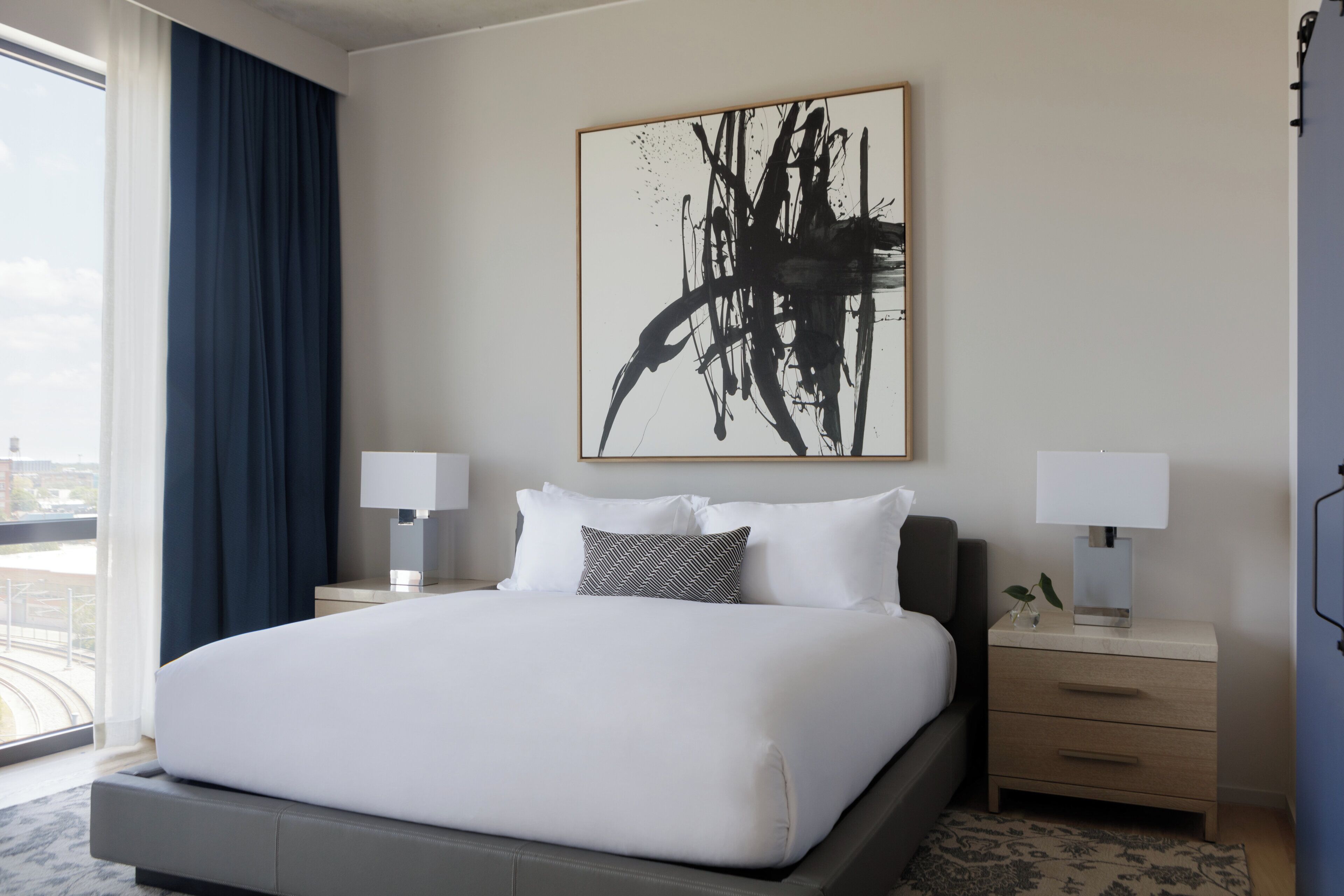 Habitación de hotel con una cama grande, dos mesitas de noche con lámparas, un cuadro abstracto de gran tamaño en la pared y vistas por la ventana.