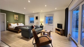 Ein modernes Wohnzimmer mit Sofa, Sessel, Fernseher und einem Fenster mit Blick auf Bäume.