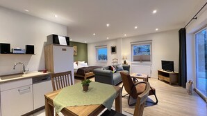 Eine moderne Wohnung mit Küche, Essbereich und Wohnzimmer. Sie verfügt über einen Kühlschrank, einen Esstisch mit Stühlen, ein Sofa, einen Fernseher und ein Fenster mit Blick.