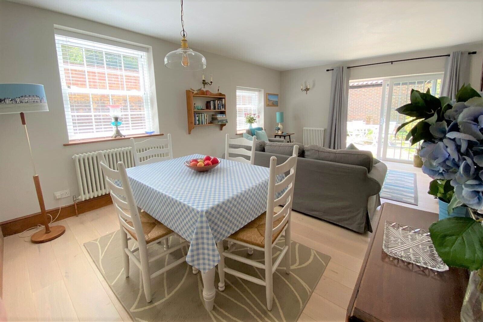 Un coin repas avec une table dressée pour quatre personnes, un canapé gris, une bibliothèque et une fenêtre avec des stores.