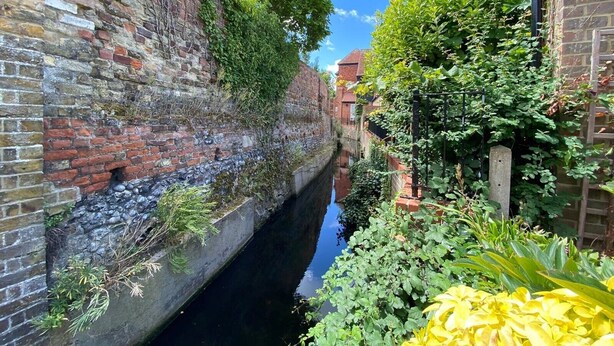 Un canal étroit bordé de murs de briques et d’une végétation luxuriante, sous un ciel dégagé.