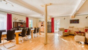 Un salon spacieux doté d’un plancher en bois, un coin repas avec une table en bois et des chaises modernes, un ensemble de canapés et une bibliothèque.