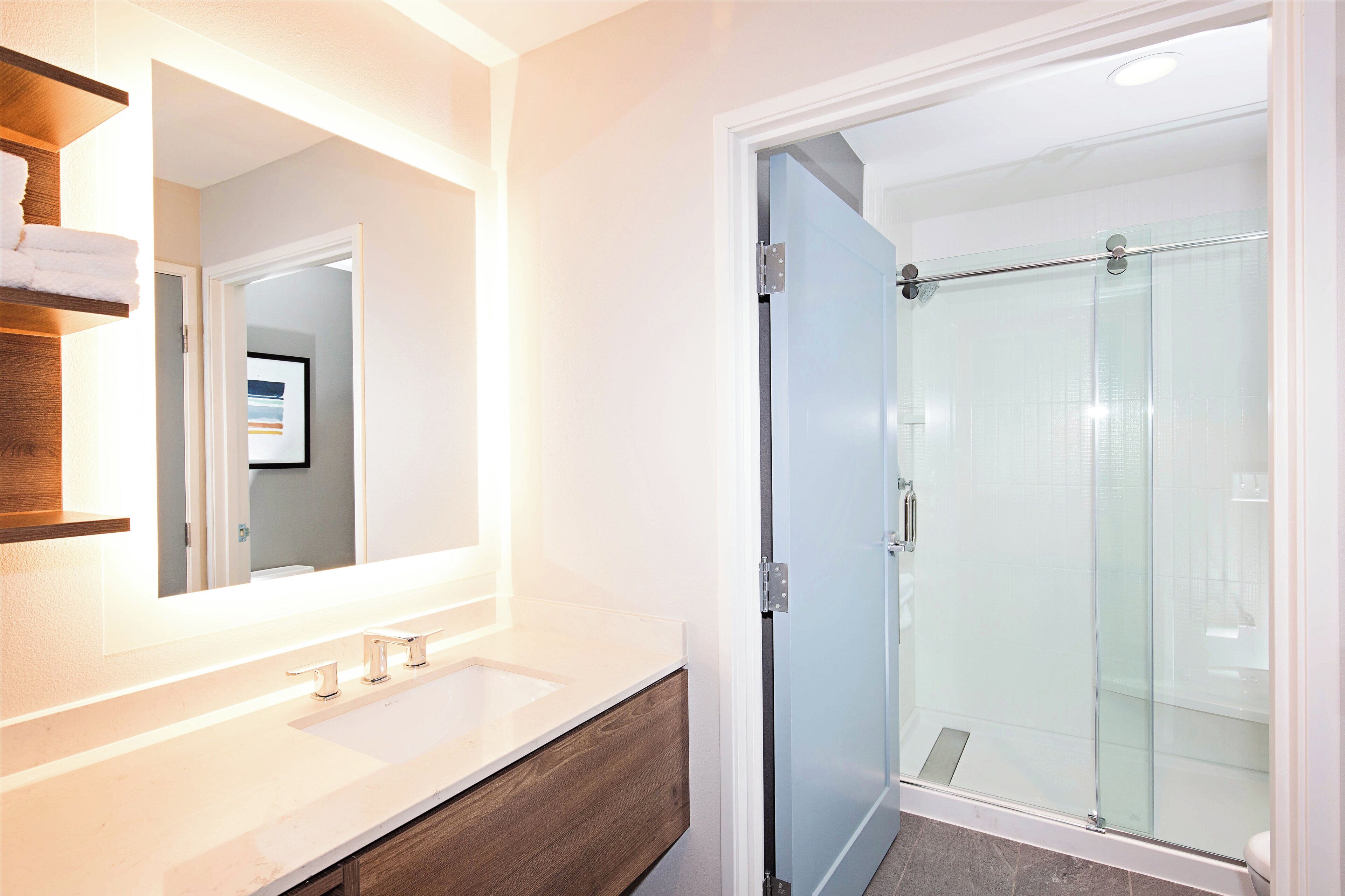 Un baño con un espejo grande, un lavabo blanco, un mueble de tocador de madera y una cabina de ducha de vidrio.