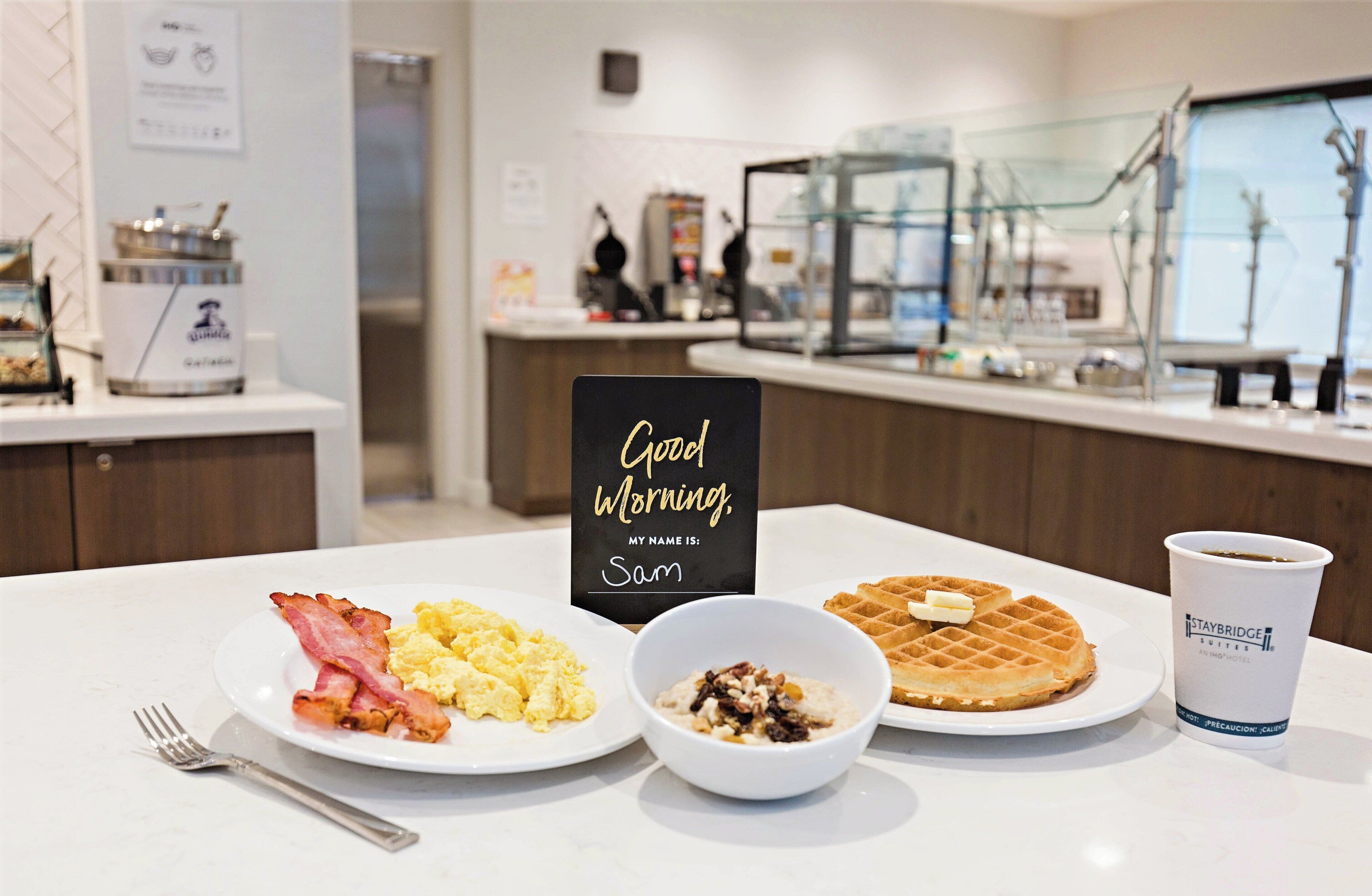 A breakfast setting with scrambled eggs, bacon, waffles, and a cup of coffee on a hotel table.