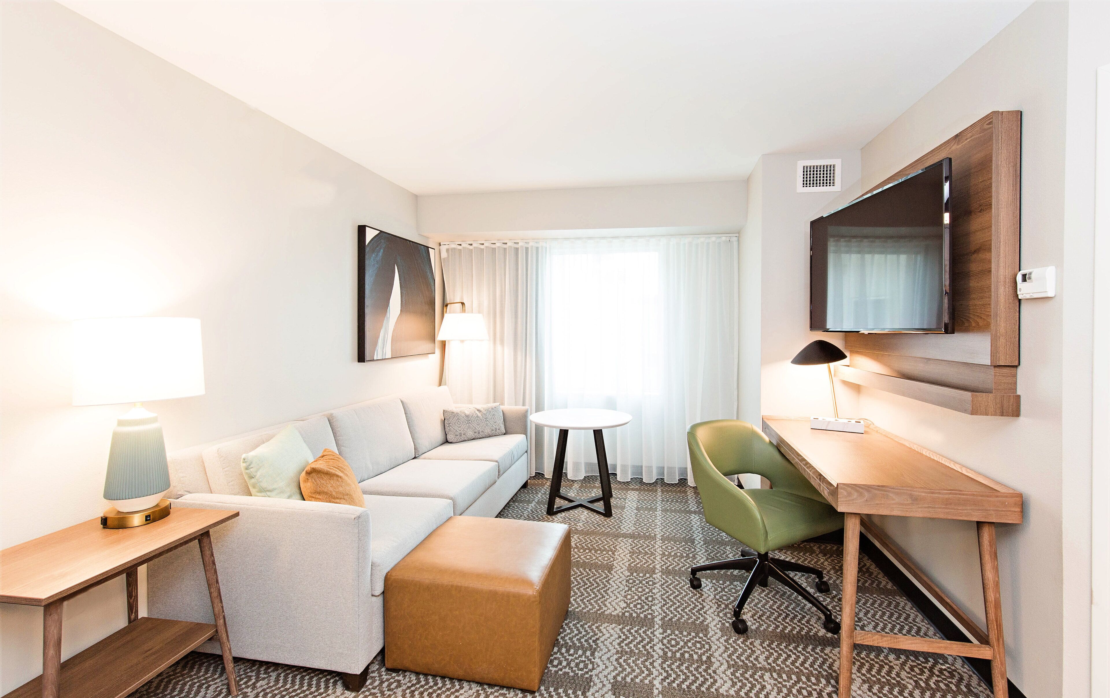 A hotel room with a sofa, a small round table, a desk with a chair, a television mounted on the wall, and a window with curtains.