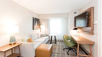 A hotel room with a sofa, a small round table, a desk with a chair, a television mounted on the wall, and a window with curtains.