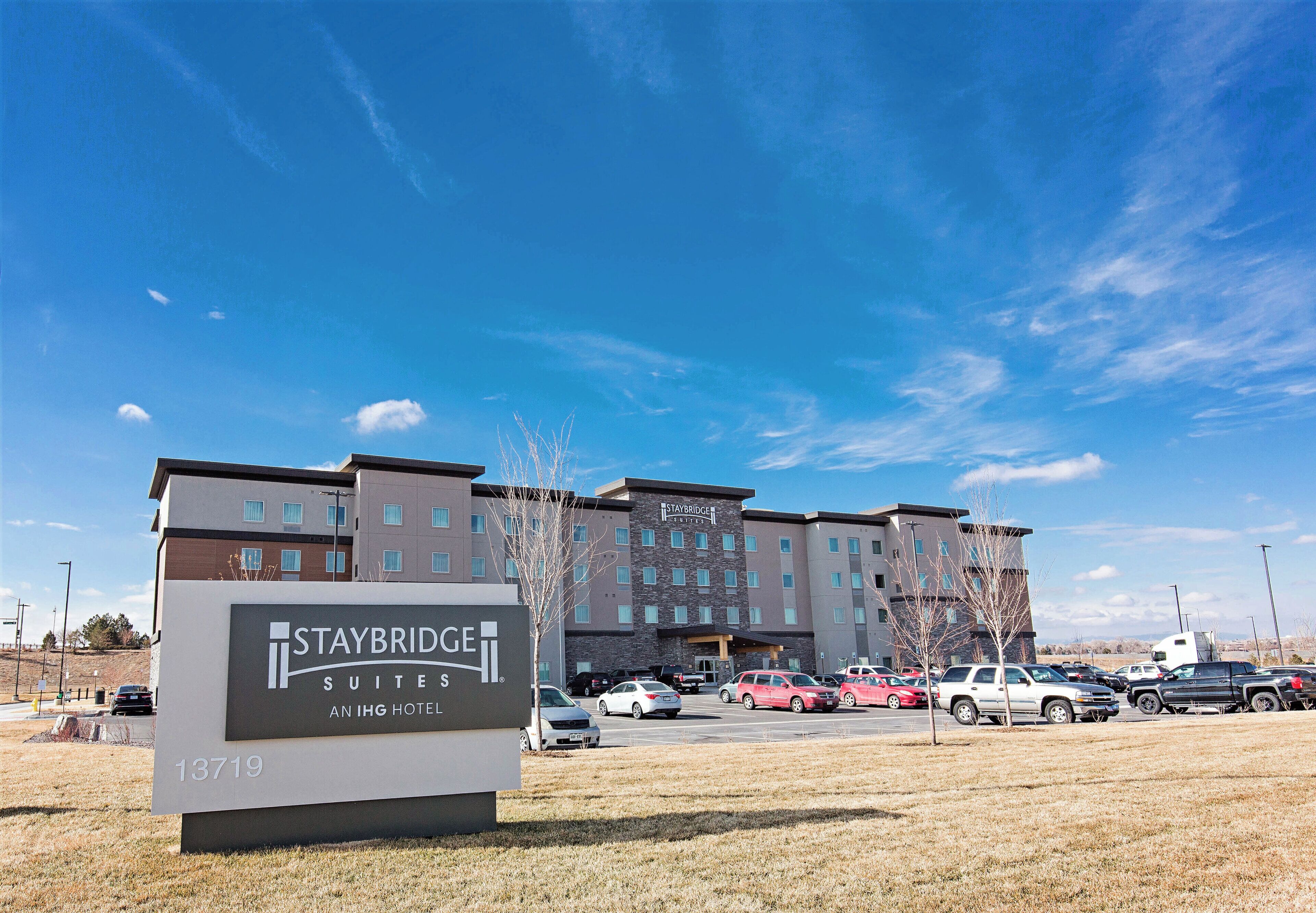 Ein Staybridge Suites Hotel mit einem Schild, das es als IHG Hotel kennzeichnet, ein Parkplatz mit mehreren Autos und einem strahlend blauen Himmel.