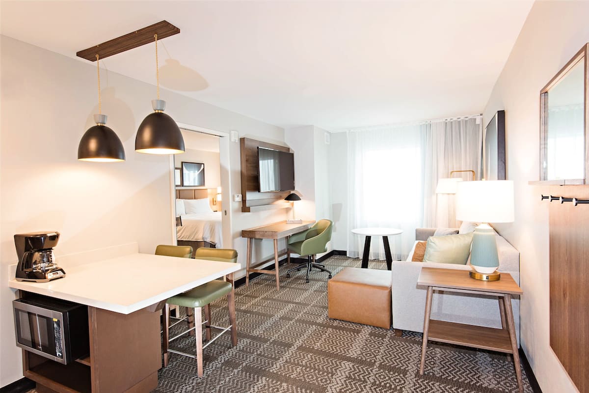A hotel room with a kitchenette, a dining area, a sofa, a TV, and a large window.
