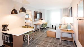 A hotel room with a kitchenette, a dining area, a sofa, a TV, and a large window.