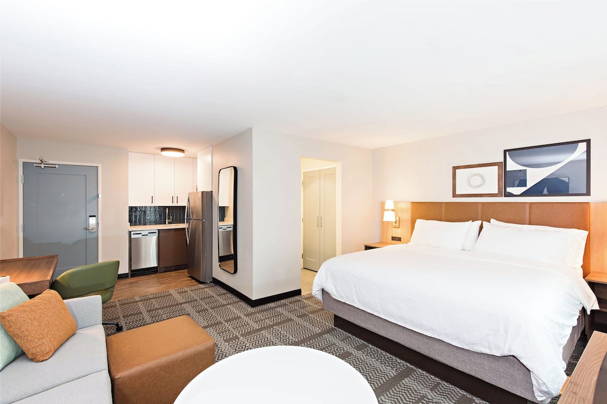 A hotel room with a large bed, a desk, a chair, a sofa, a small round table, and a kitchenette.
