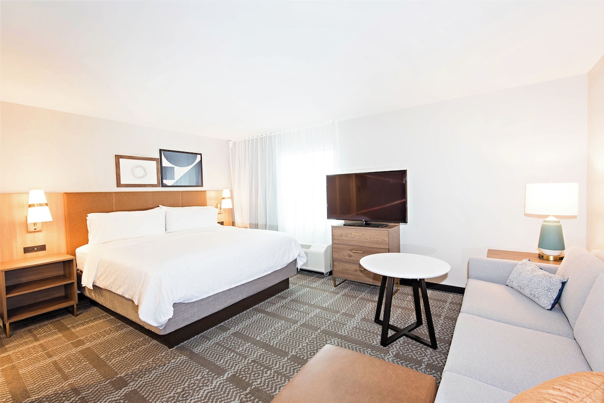 A hotel room with a bed, a TV, a sofa, a small table, and a nightstand with a lamp.