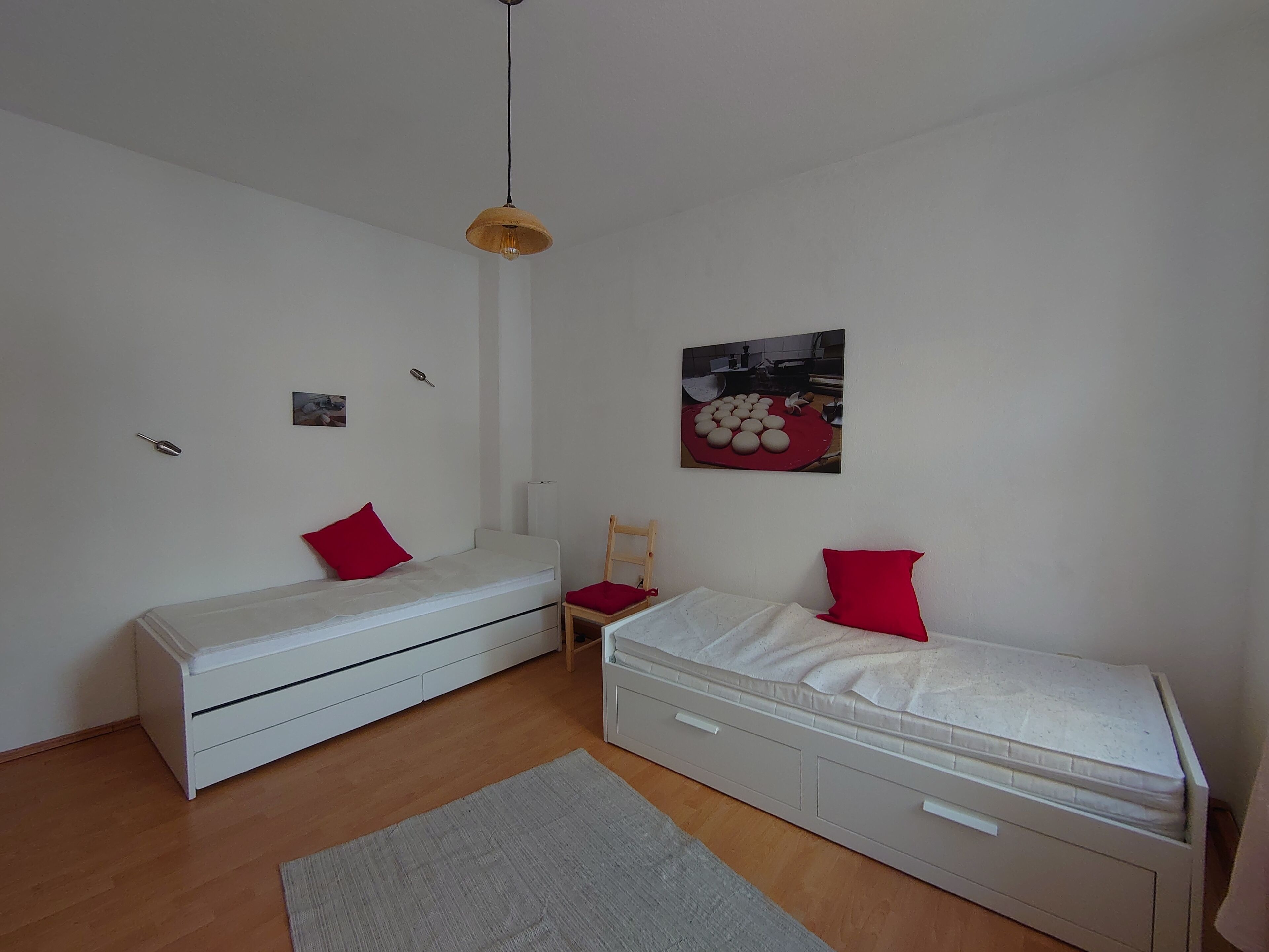 A small, clean room with two single beds, a wooden chair, a small table, and a hanging light fixture.