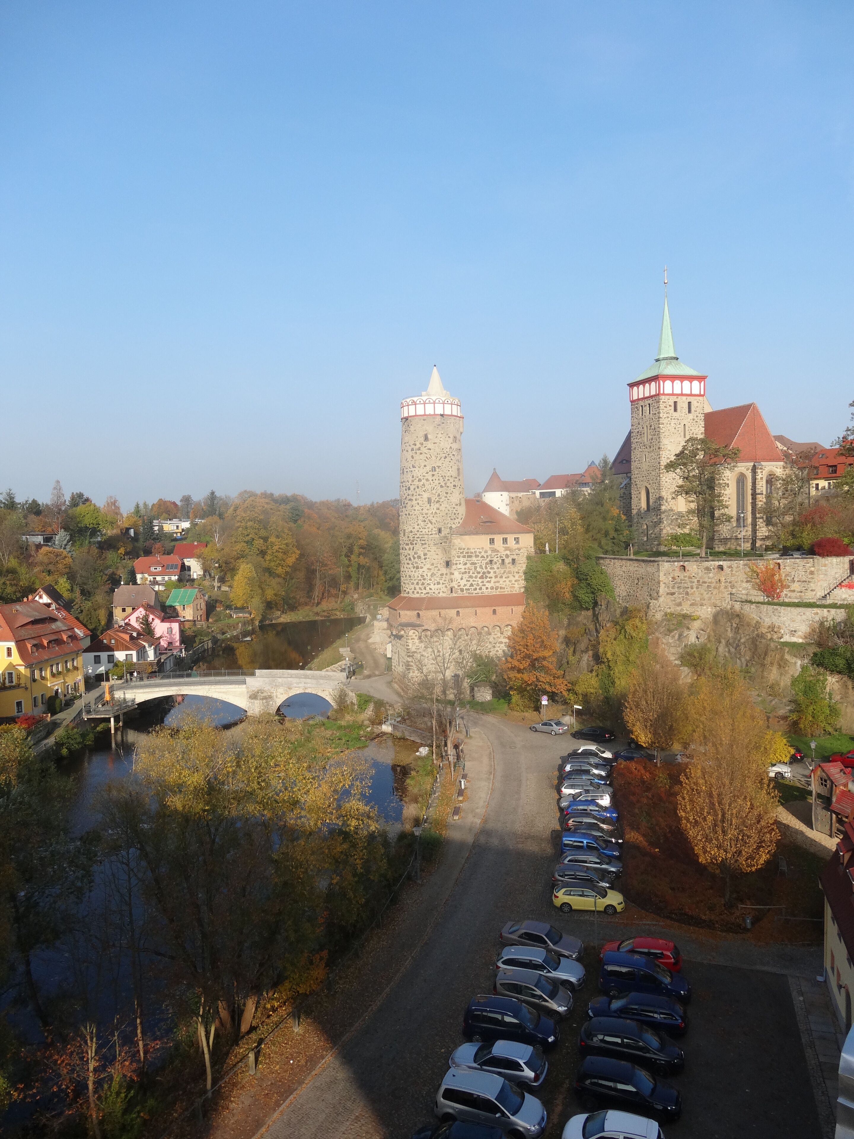 A historic stone castle with a prominent tower and a church with a green spire. A bridge crosses a river in the foreground, and there is a parking area with cars.