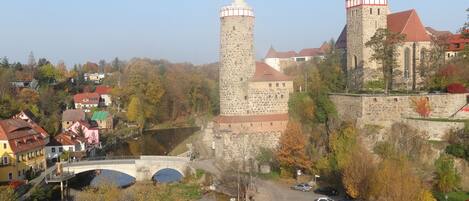 A historic stone castle with a prominent tower and a church with a green spire. A bridge crosses a river in the foreground, and there is a parking area with cars.