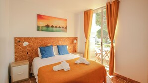 A hotel room with a bed, a nightstand, a painting of a sunset over water, and a view of a balcony with orange curtains. - Pé na Areia (Castro Marim)