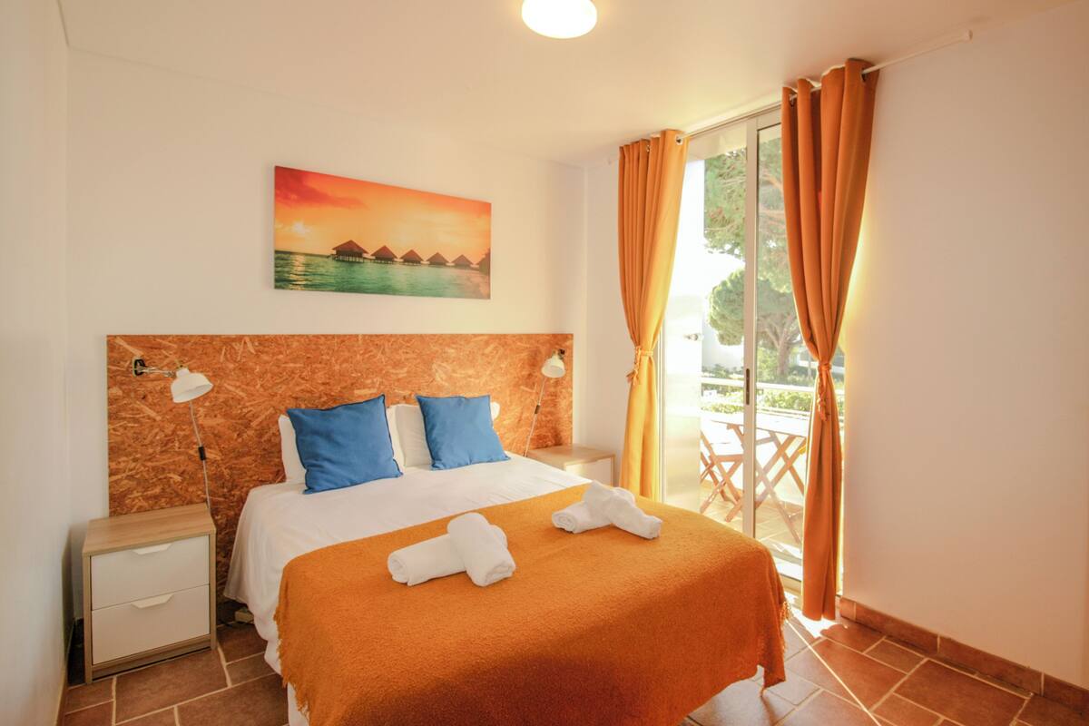 A hotel room with a bed, a nightstand, a painting of a sunset over water, and a view of a balcony with orange curtains.
