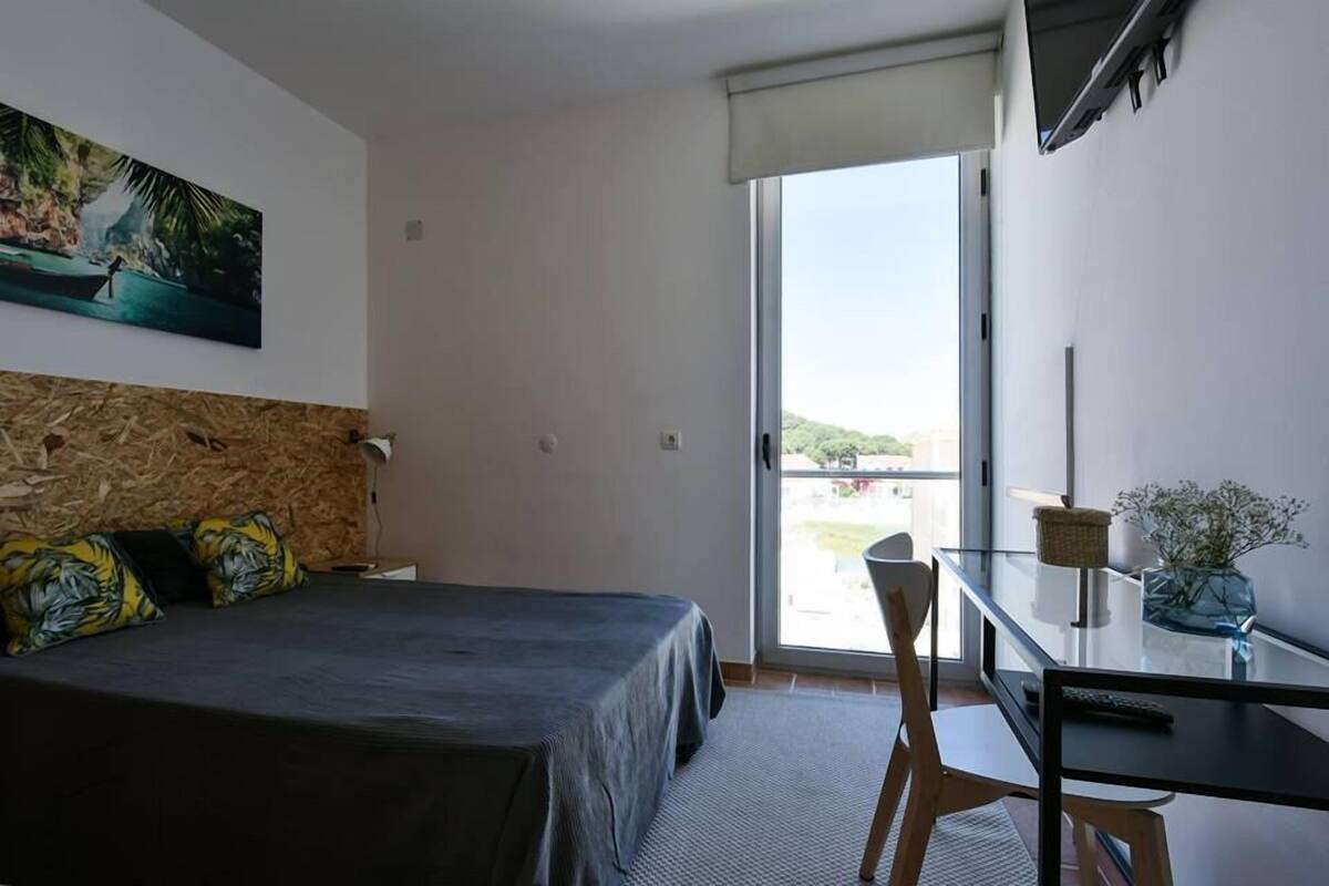 A hotel room with a bed, a desk, a chair, a window, and a painting on the wall.