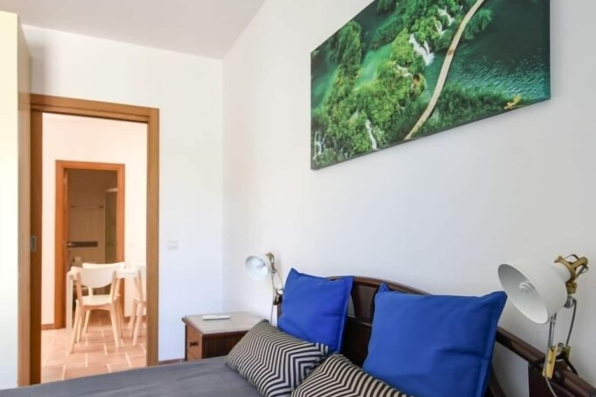 A bedroom with a bed, a desk, a chair, and a painting on the wall.
