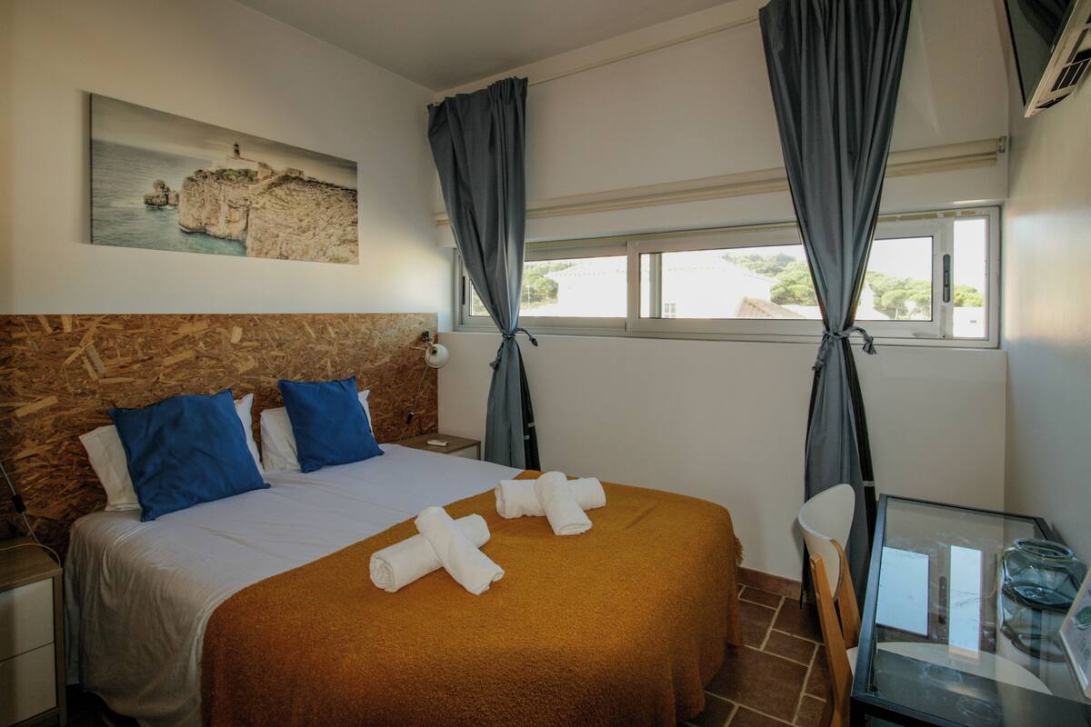 A hotel room with a bed, a painting on the wall, a window with curtains, a bedside table, and a chair.