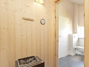 A wooden sauna room with a sauna thermometer, a wooden bench, and a window leading to a bathroom with a toilet. - Holiday Home in Großenbrode (Grossenbrode)