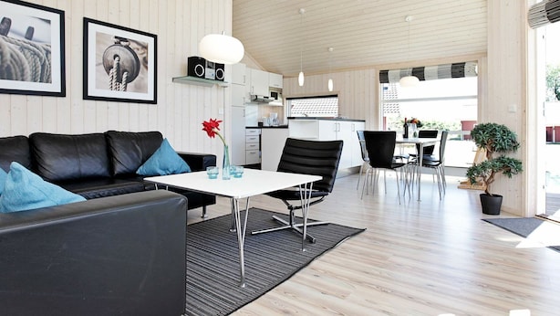 A modern living room with a black leather sofa, a white coffee table, and a dining area with a white table and black chairs. There are framed pictures on the wall, a kitchen in the background, and a wooden ceiling. - Holiday Home in Großenbrode (Grossenbrode)