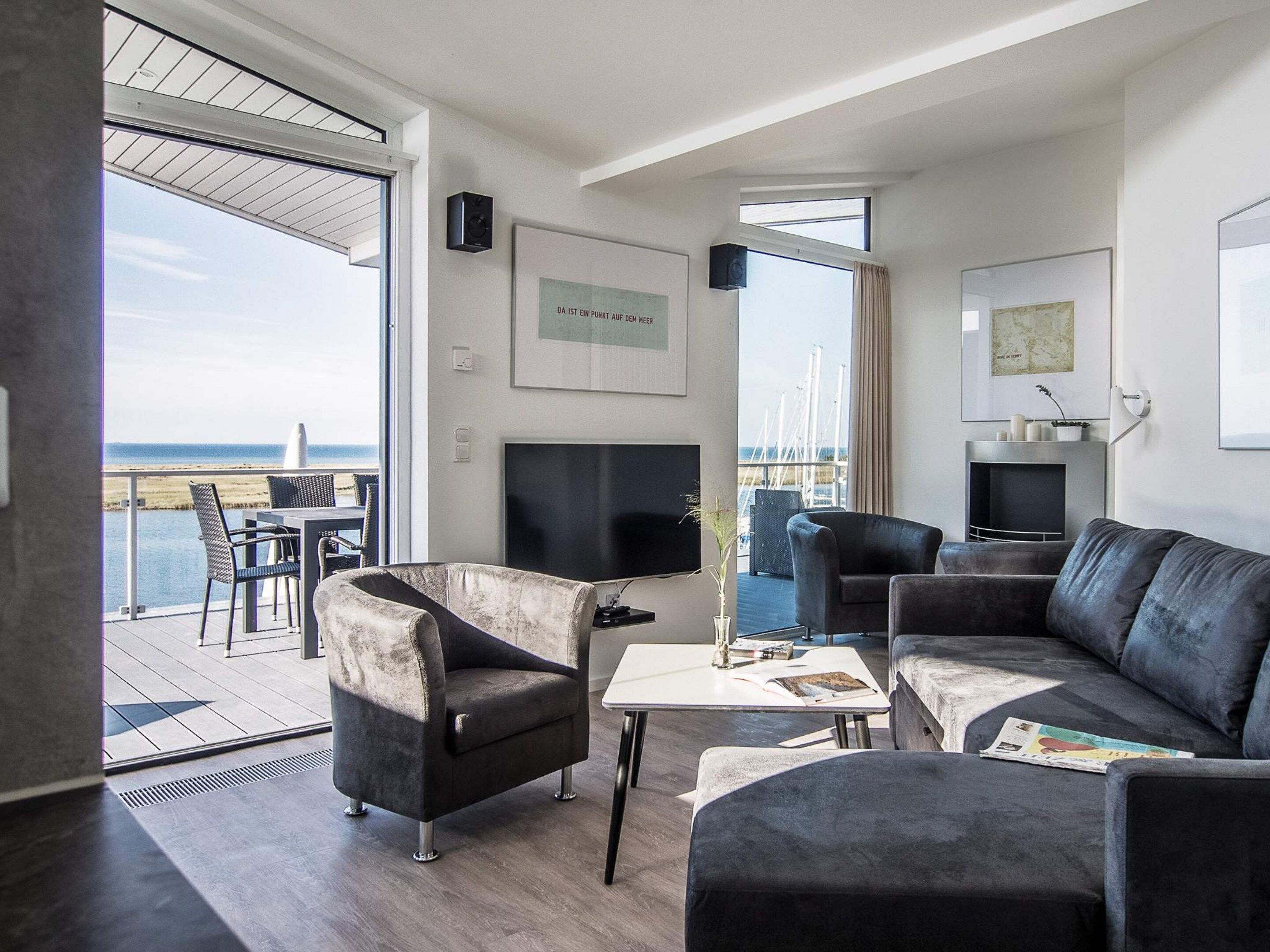 A modern living room with a large TV, comfortable seating, and a view of the sea.