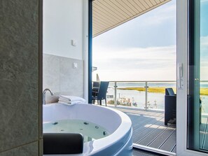 Ein modernes Badezimmer mit einer freistehenden Badewanne, einer Duschkabine aus Glas und Blick auf einen Yachthafen mit Booten und einem Strand.
