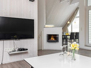 A modern room with a flat-screen TV mounted on a white wall, a fireplace with a visible fire, a dining area with a table and chairs, and a large window allowing natural light. - Holiday Home in Wendtorf (Wendtorf)
