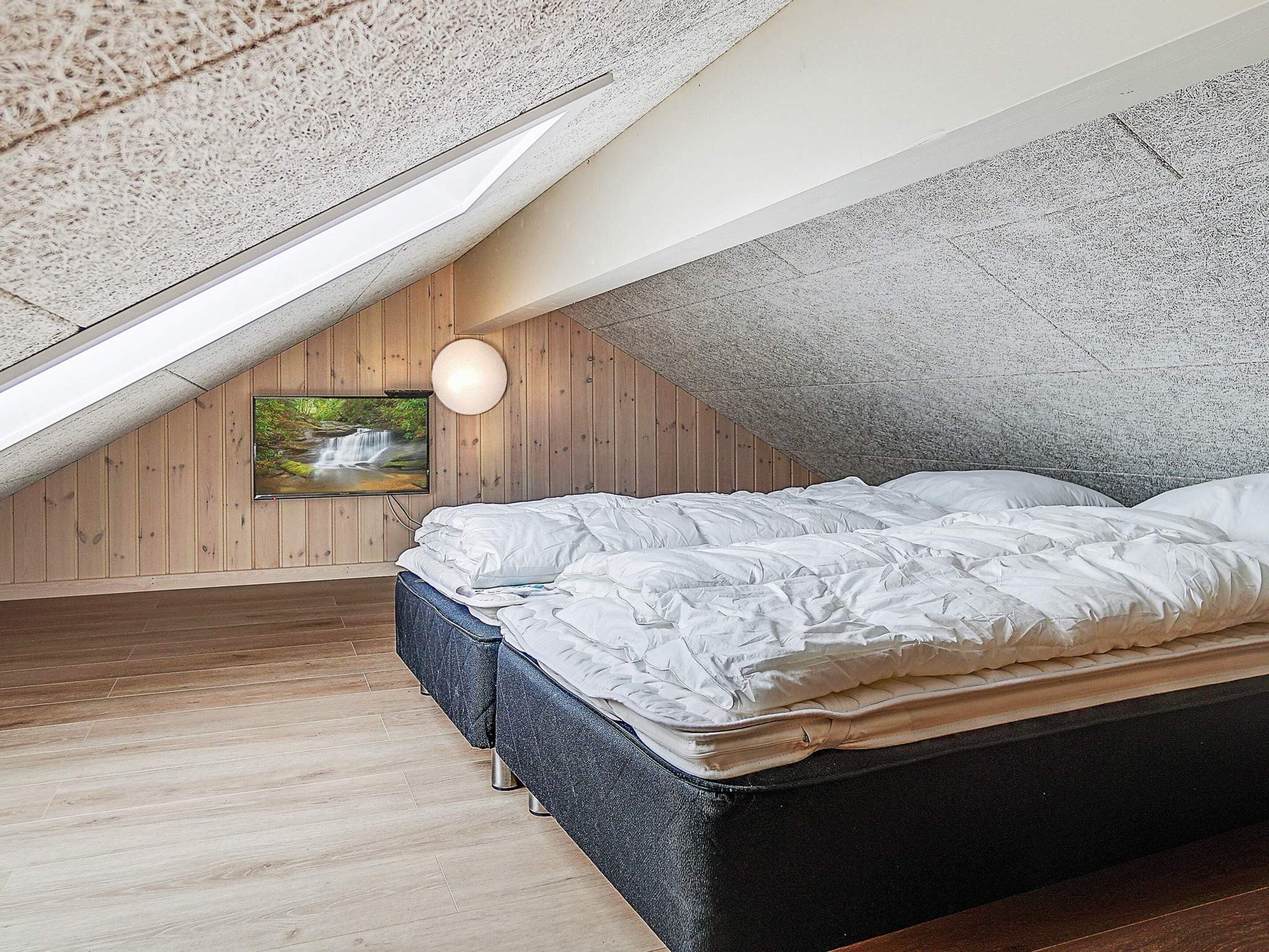 A bedroom with a sloped ceiling, a bed with white bedding, a mounted TV, and a wooden floor.