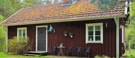 A small, single-story building with a tiled roof, a front door, and a small porch with chairs.