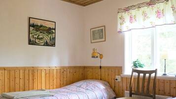 A room with a wooden bunk bed, a single chair, a small table, a wall-mounted lamp, and a window with floral curtains.