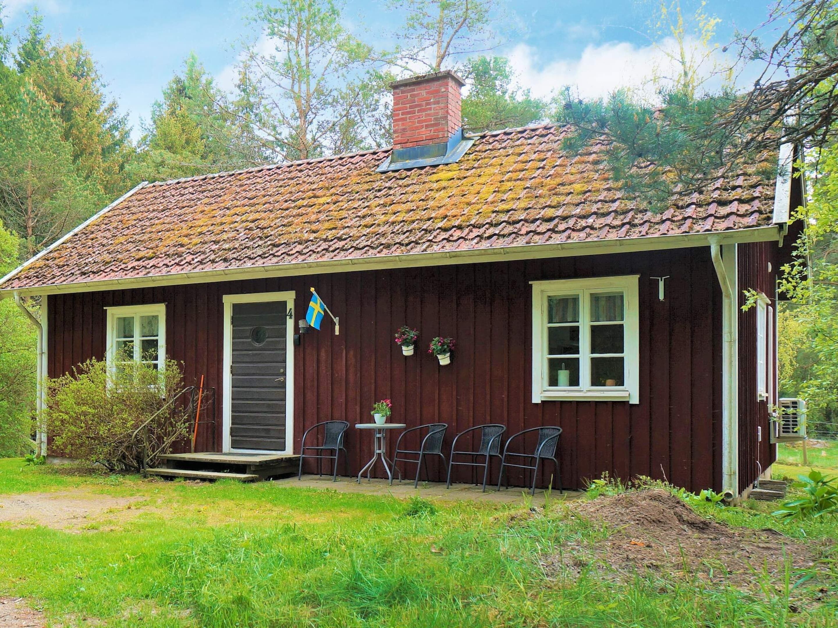 A small, single-story wooden house with a tiled roof, a front door, and a small porch with four chairs.