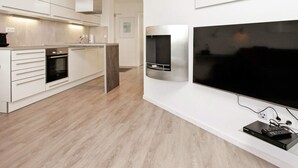 A modern kitchen with white cabinetry, a built-in oven, a stainless steel range hood, and a large flat-screen TV mounted on the wall.