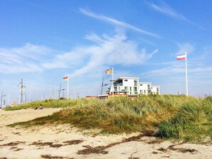 A coastal building with a flat roof, surrounded by sand and grass, with flags in the background. - Holiday Home in Wendtorf (Wendtorf)
