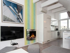 A modern living room with a flat-screen TV, a fireplace, and a kitchen area with white cabinets and a window.