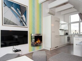Una sala de estar moderna con televisor de pantalla plana, chimenea y área de cocina con muebles blancos y una ventana.