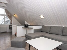 Un espacio habitable moderno estilo loft con techo inclinado, un sofá modular, una mesita blanca y una zona de kitchenette.