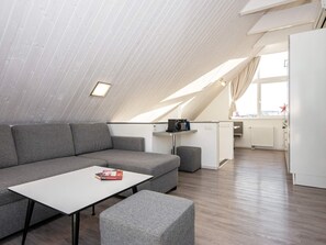 A modern loft apartment with a sloped ceiling, a grey sofa, a white table, and a kitchenette area. - Holiday Home in Wendtorf (Wendtorf)