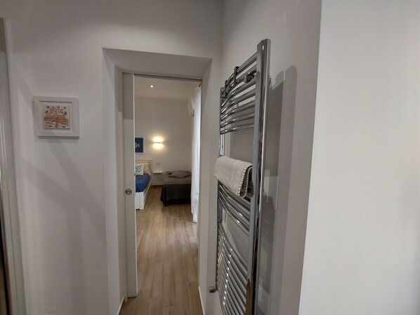 A narrow hallway with a metal towel rack on the right and a door leading to a bedroom with a bed and a nightstand.
