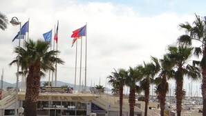 A building with multiple flags, palm trees, and a marina in the background. - Central 2BR with Balcony & A/C - 3rd Floor - Cavalaire-sur-Mer (Cavalaire-sur-Mer)