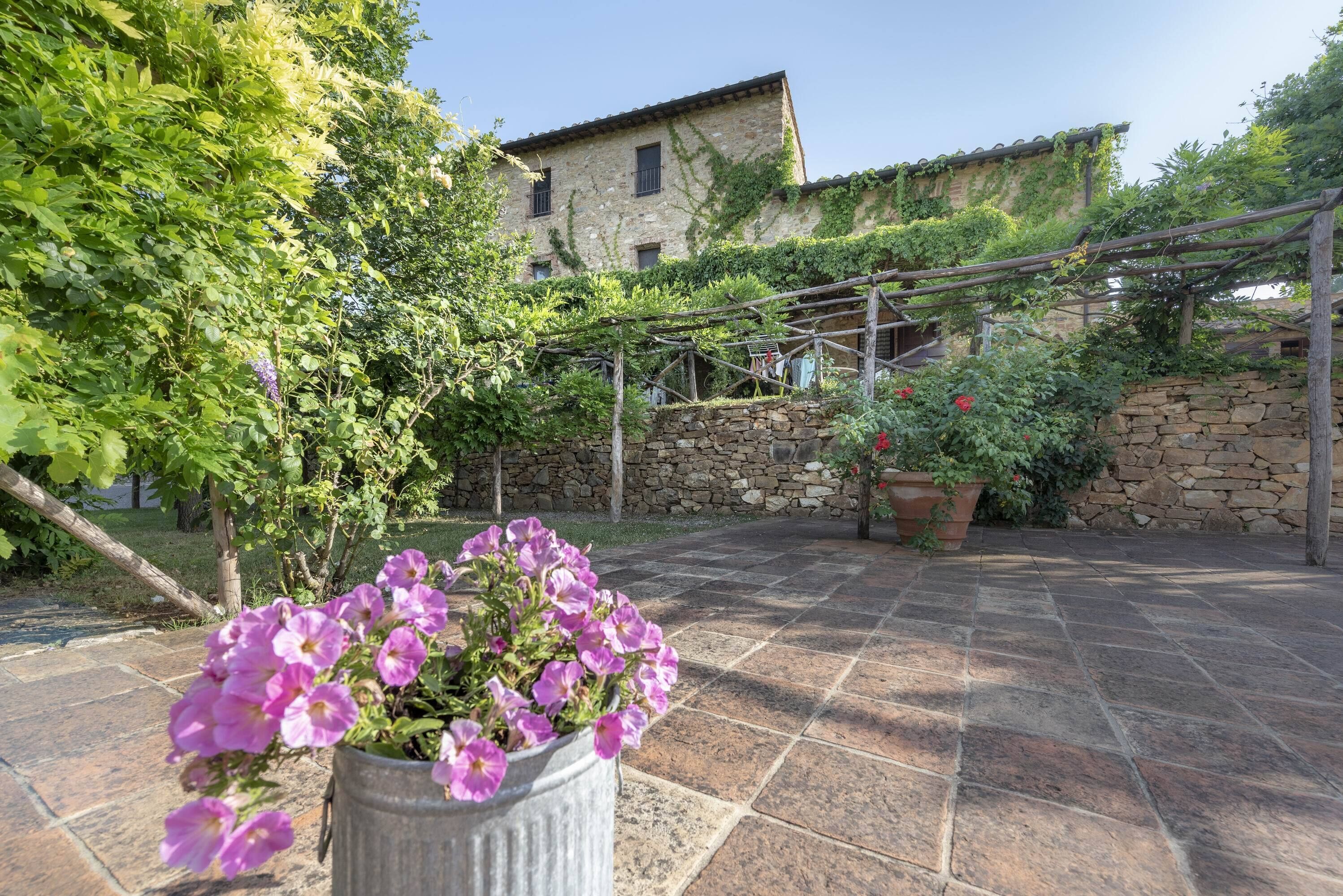 A stone building with a garden area featuring a potted plant with purple flowers and a pergola.